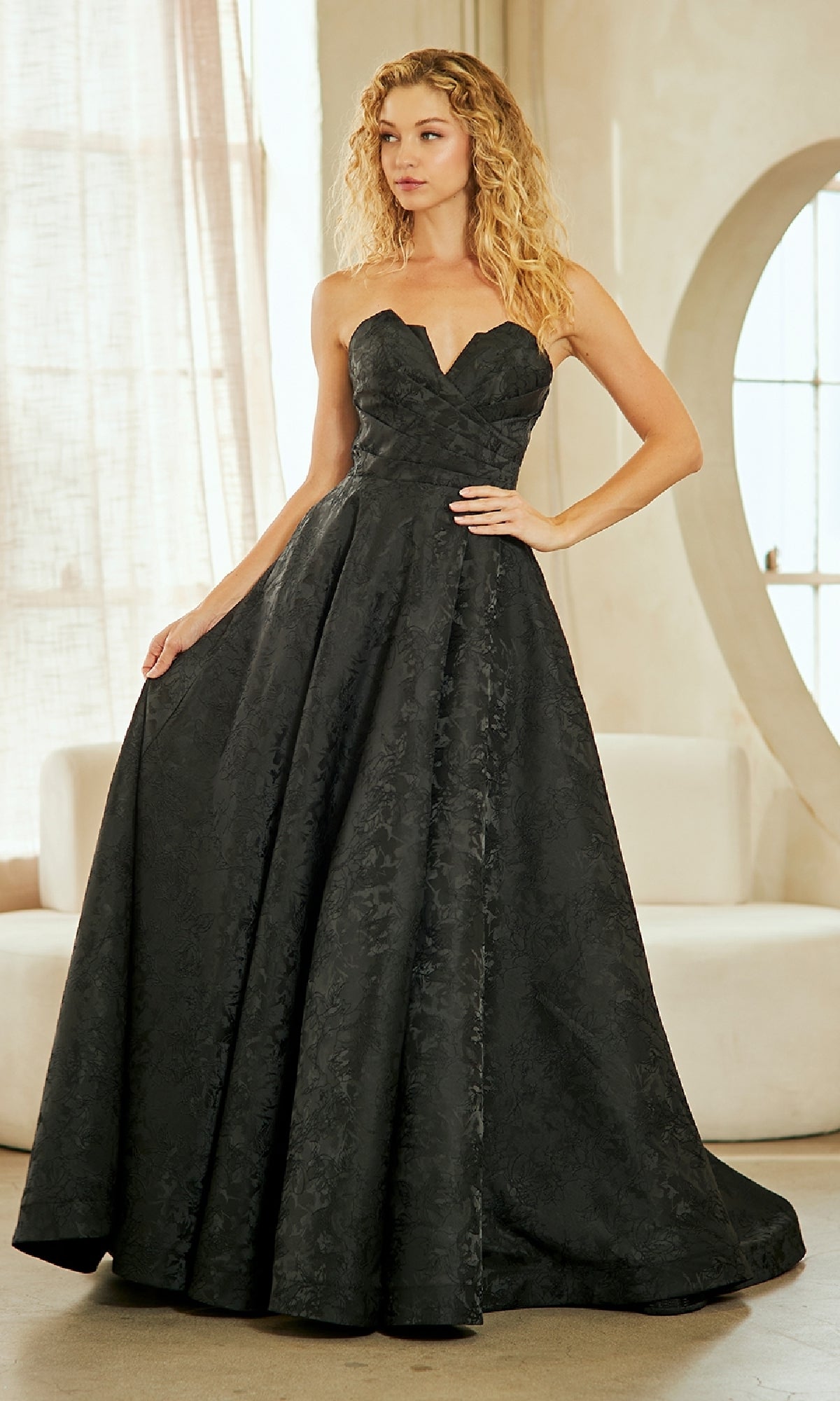 A woman in an Amelia Couture 8061 Textured Strapless Prom Gown stands indoors by a window.